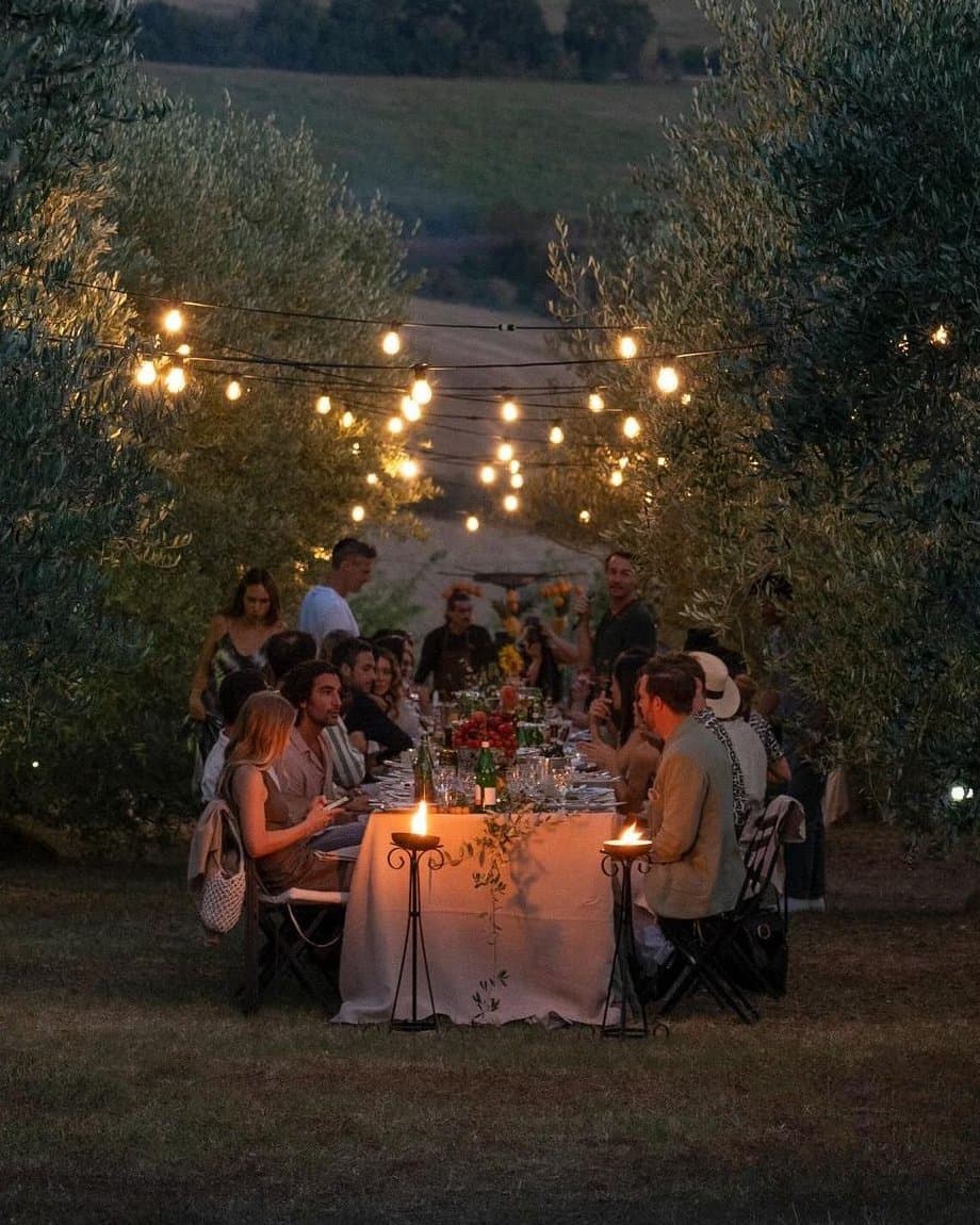 Cena larga al aire libre bajo luces colgantes, entre árboles (foto ilustrativa)